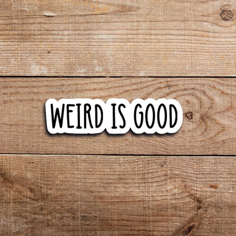 May include: A white sticker with the phrase "WEIRD IS GOOD" in black, sans-serif font. The sticker is rectangular with rounded corners and is set against a wooden background with horizontal planks.