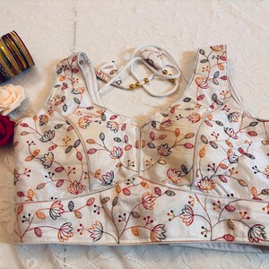 May include: A sleeveless white blouse featuring an intricate floral embroidery pattern in shades of orange, red, and dark blue. The garment includes a sweetheart neckline, a wide waistband, and white tie-up straps with gold beads. A stack of colorful bangles and two fabric roses are placed nearby.