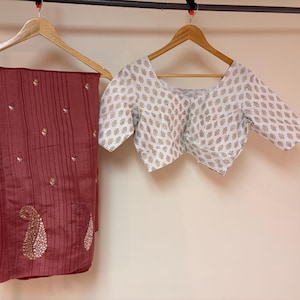 May include: A maroon saree with vertical stripes and gold paisley designs, hanging on a wooden hanger. A white blouse with a beige leaf pattern is also on a wooden hanger. The saree has tassels.