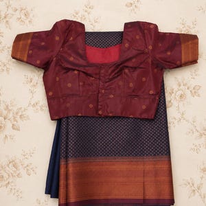 May include: A maroon blouse with gold polka dots and a square neckline is displayed over a navy blue saree with a gold and maroon border. The saree has a repeating pattern. The background is a floral wallpaper.