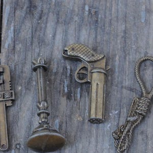 May include: A selection of small, antique brass objects, including a poison bottle, dagger, wrench, candlestick, pistol, rope, nail, and horseshoe. The objects are arranged on a wooden surface next to a ruler.