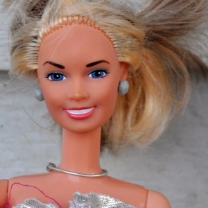 May include: A vintage doll with blonde hair styled in a ponytail. The doll has blue eyes, a silver dress, and a silver necklace. A wooden ruler with inch measurements is visible in the background.