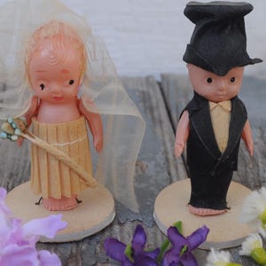 Peut inclure: Deux figurines de gâteau de mariage vintage. La mariée porte un voile et tient un bâton avec de petites fleurs. Le marié porte un smoking noir et un haut de forme. Les poupées sont sur des socles ronds, avec des fleurs éparpillées.