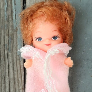 May include: A small vintage doll with red hair and blue eyes. The doll is wearing a pink robe with white lace trim. The doll's arms are outstretched. The background is a weathered, blue-gray wooden surface.