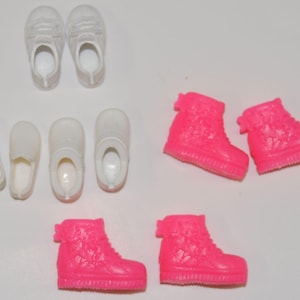 Könnte beinhalten: Eine Sammlung von Puppenschuhen in verschiedenen Stilen und Farben. Es gibt zwei Paar weiße Turnschuhe, drei Paar weiße Slipper und drei Paar leuchtend pinkfarbene High-Top-Sneaker. Am unteren Rand des Bildes befindet sich ein Holzlineal.