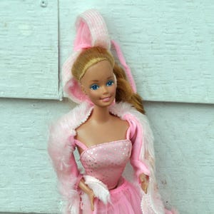 Op de afbeelding: Een vintage Barbie-pop met een roze jurk met een doorschijnende rok en een bijpassende roze jas met bont. De pop heeft blond haar en blauwe ogen. De outfit bevat een bijpassende capuchon.
