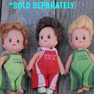 May include: Three vintage doll figures with different hair colors. Two dolls wear green overalls with "LILY LIME" text, and one doll wears red overalls with "DOLLY COLA" text. The dolls have painted faces and are posed together.