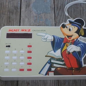 Puede incluir: Calculadora vintage de Mickey Mouse con cuerpo blanco y un gráfico colorido de Mickey Mouse con toga de graduación. La calculadora tiene una pantalla digital y botones numéricos. Una cuerda blanca está unida en la parte superior.