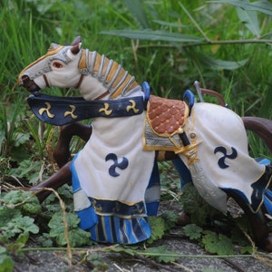 May include: A detailed figurine of a knight's horse, featuring a white body with blue and gold accents. The horse has a brown mane and tail, and is adorned with a saddle and a decorative cloth with a stylized symbol. The figurine is set against a backdrop of green foliage.