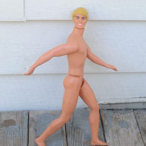 May include: A vintage doll with a tan plastic body and blonde hair. The doll is posed in a running stance with arms outstretched. The doll is standing on a weathered wooden surface.