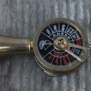 May include: A brass ship's telegraph key chain. The key chain has a circular dial with the words "AHEAD" and "ASTERN" and a small sailboat graphic. The key chain is on a weathered wooden surface.