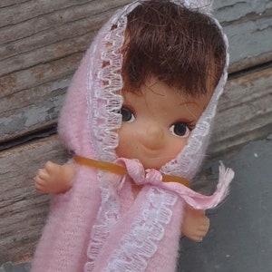 Könnte beinhalten: Eine kleine rosafarbene Puppe, die ein weißes, mit Spitze besetztes, kapuzenförmiges Kleid trägt. Die Puppe hat braunes Haar und blaue Augen.