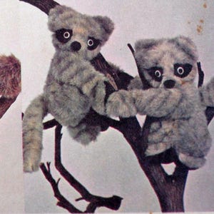 May include: A collection of soft toy animals, including monkeys, koalas, and raccoons, arranged on tree branches. The toys are in shades of brown, grey, and black, with detailed features and outstretched limbs.