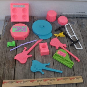 May include: A collection of colorful toy musical instruments, including pink guitars, a green piano, and a blue drum set. The set also includes a pink speaker, drumsticks, and a blue disc. The text "NEW KIDS ON THE BLOCK" is visible on some pieces.
