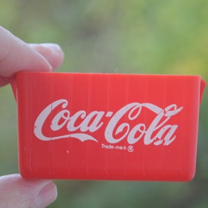 Puede incluir: Contenedor de plástico rojo con el logotipo de Coca-Cola en letras blancas y las palabras "Trade-mark" y un símbolo de marca registrada.
