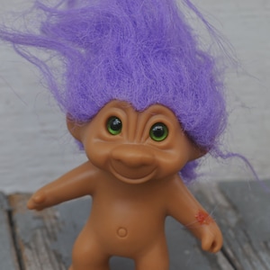 May include: A purple haired troll doll with green eyes and a smiling face. The doll is made of plastic and has a brown body.