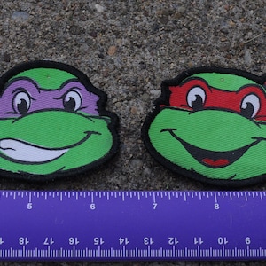 May include: Two embroidered patches featuring cartoon turtle faces with green skin and black outlines. One turtle wears a purple mask, the other a red mask. A purple ruler is positioned below the patches, showing they are approximately 3 inches wide.