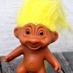 May include: A small, brown troll doll with yellow yarn hair. The troll has large, round, orange eyes and a wide smile.