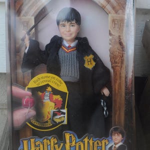 May include: A Harry Potter doll in a clear plastic package. The doll is wearing a black robe with a Gryffindor crest and holding a wand. The packaging includes the text "Harry Potter and the Sorcerer's Stone" and "Hogwarts Heroes Harry".
