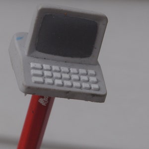 Op de afbeelding: Een grijze plastic speelgoedlaptop met een wit toetsenbord en een zwart scherm. De laptop is bevestigd aan een rood potlood.