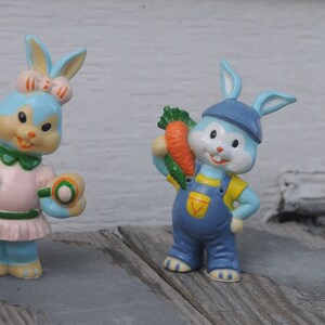 May include: Two blue bunny figurines, one wearing a pink dress and holding a ball, the other wearing overalls and holding a carrot.