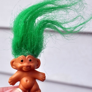 May include: A small plastic troll doll with green hair and a brown body. The troll is smiling and has large eyes.