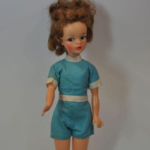 May include: A vintage doll with auburn hair and blue eyes, wearing a turquoise playsuit with white trim. The doll has rosy cheeks and red lips, and is posed against a plain background. The doll's outfit includes a matching belt.