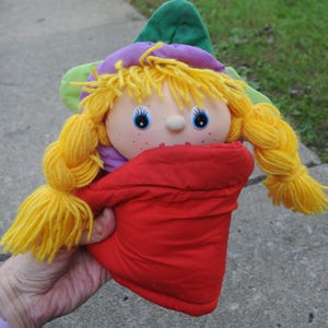 May include: A plush doll with yellow yarn hair styled in two braids, blue eyes, and freckles. The doll wears a purple and green hat and is wrapped in a solid red fabric pouch that covers the lower body, leaving the head and arms visible.