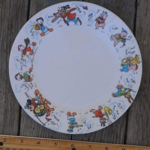 May include: A white ceramic plate with a blue and white border featuring a cartoon band of Disney characters playing musical instruments. The characters include Mickey Mouse, Donald Duck, Goofy, and Pluto.