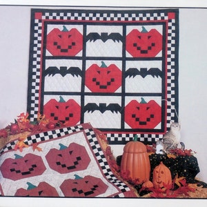 Peut inclure: Une courtepointe sur le thème d'Halloween avec des citrouilles rouges et des chauves-souris noires sur fond blanc, encadrée d'une bordure à carreaux noirs et blancs. Une plus petite courtepointe avec des motifs de citrouilles similaires est également visible, ainsi que des citrouilles décoratives et des feuilles d'automne.