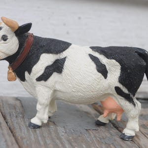 May include: A figurine of a Holstein cow, featuring a black and white spotted pattern. The cow has a brown collar with a gold bell, and tan horns. The udder is pink. The figurine is standing on a wooden surface.