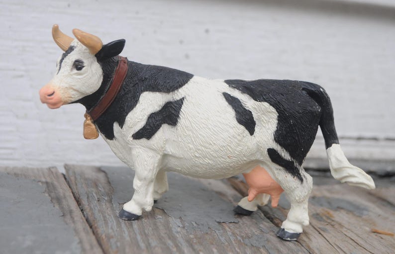 May include: A figurine of a Holstein cow, featuring a black and white spotted pattern. The cow has a brown collar with a gold bell, and tan horns. The udder is pink. The figurine is standing on a wooden surface.