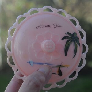 May include: Pink plastic trinket box with a white lace trim. The box has a painted design of a palm tree and a flower. The text "Miami, Fla." is written on the box.