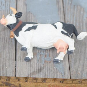 May include: A small, decorative cow figurine with a black and white pattern. The cow has a brown collar, a gold bell, and small horns. The udder is pink. The figurine is on a weathered wooden surface.