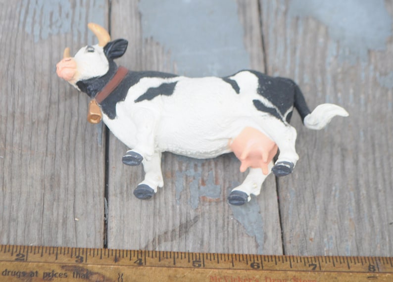 May include: A small, decorative cow figurine with a black and white pattern. The cow has a brown collar, a gold bell, and small horns. The udder is pink. The figurine is on a weathered wooden surface.