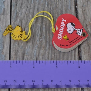 May include: A red heart-shaped book with the word "SNOOPY" printed on the front. The book is attached to a yellow string with a yellow plastic charm of Snoopy's bird friend Woodstock.