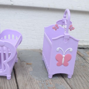 May include: Two lavender toy cribs. One has a heart-shaped headboard, the other has a pink butterfly applique. The second crib has a mobile with pink butterfly accents. Both cribs have a slatted design and are made of plastic.
