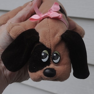 May include: A brown and black plush toy dog with large, black eyes and a pink bow. The dog is wearing a pink bow around its neck.