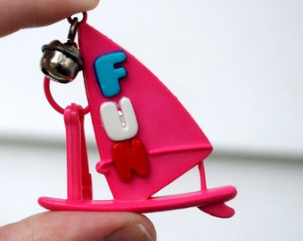 May include: A pink windsurfing keychain with the word "FUN" in blue, white, and red letters. The keychain has a small silver bell attached to the top. The windsurfing board and sail are made of plastic.
