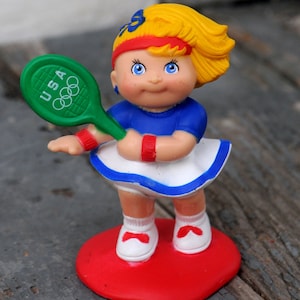 May include: A plastic figurine of a girl wearing a blue and white tennis outfit, holding a green tennis racket with the word "USA" and Olympic rings on it. She is standing on a red base.