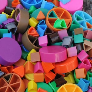 May include: A colorful assortment of plastic toy pieces in various shapes and colors. The image features a mix of geometric shapes, including circles, triangles, and cubes, in shades of pink, orange, green, blue, and brown. The pieces appear to be from a children's toy set.