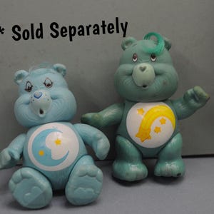 May include: Two Care Bear plush toys. The Care Bear on the left is blue with a yellow crescent moon and stars on its belly. The Care Bear on the right is green with a yellow shooting star and stars on its belly. *Sold Separately*