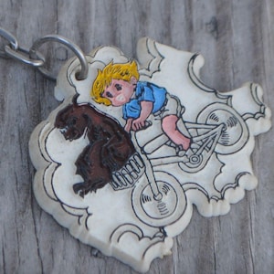 May include: A white metal keychain with a cartoon image of a boy riding a bicycle with a brown dog running beside him. The boy is wearing a blue shirt and red shorts. The keychain has a silver chain.