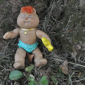 May include: A vintage Cabbage Patch Kids doll in a seated position. The doll has brown skin, a round face, and orange hair. It wears a gold necklace, blue diaper with white polka dots, and holds a yellow object. The doll is set against a natural background.