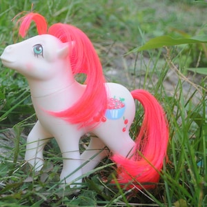Könnte beinhalten: Ein weißes Pony mit pinkfarbenem Haar und einem Kirschmuster an der Seite. Das Pony steht in einem Stück grünem Gras.
