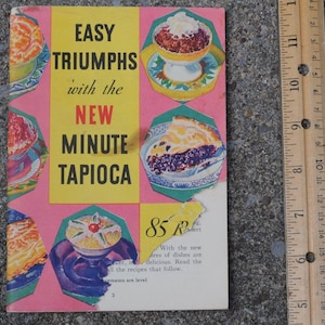 May include: Vintage cookbook cover with the title "EASY TRIUMPHS with the NEW MINUTE TAPIOCA." The cover is pink and yellow with illustrations of desserts. A wooden ruler is on the right side of the image, showing measurements in inches.