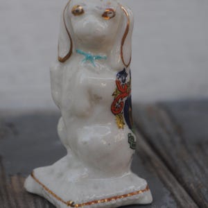 May include: A small, white ceramic dog figurine with gold accents on the ears and base. The dog has a blue bow and a colorful emblem on its side. The figurine is standing on a square base.