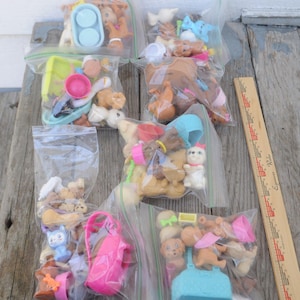 May include: A collection of small plastic toy puppies and accessories, including bowls, hats, and purses, are contained in clear plastic bags. The toys are various shades of brown, white, and blue. A wooden ruler is on the right side of the image.