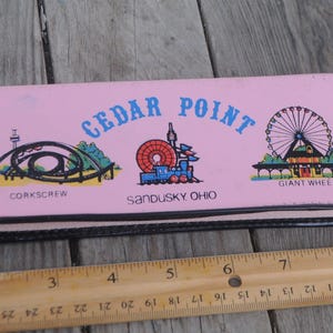 Może przedstawiać: Różowy portfel z parku rozrywki Cedar Point z obrazkami olbrzymiego koła, kolejki górskiej Corkscrew i pociągu. Portfel ma czarny zamek błyskawiczny, a na przodzie nadrukowany jest tekst "Cedar Point, Sandusky Ohio".