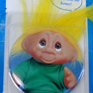 May include: A yellow-haired troll doll with a green shirt and a blue background. The troll is smiling and has a speech bubble above its head that says "Please take me home!"